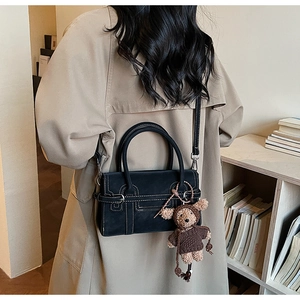 Black matte handbag with white stitching and bear charm shown on a khaki trench coat