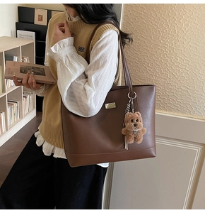 Detail of a brown leather tote bag featuring a silver plaque and cute teddy bear charm