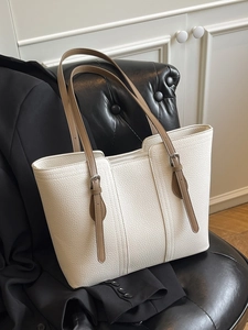 Minimalist off-white litchi grain tote bag with brown straps on a black leather sofa