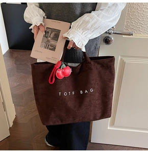 Detail of a brown suede tote bag with a red cherry charm and white logo text
