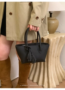 Detail of black pebbled leather tote bag with dual tassels held by a model in a trench coat