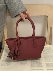 Chic burgundy faux leather tote bag with elegant tassels held against a grey coat