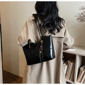 Detail of glossy black faux leather tote bag with tassel charm worn over khaki trench coat