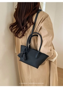 Detail of a black bucket bag with double tassels and thin shoulder strap over a beige coat