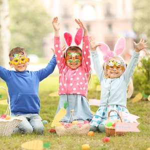 Drei Kinder tragen bunte Oster-Partybrillen aus Papier auf einer sonnigen Rasenfläche