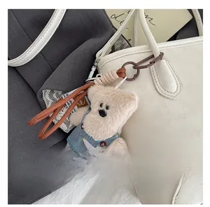 Detail of plush bear bag charm in denim overalls hanging on a white leather handbag
