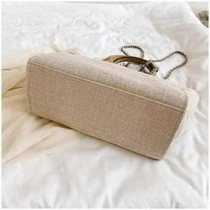 Detalle de bolso tote tejido beige claro con correa de cadena plateada sobre sábana blanca