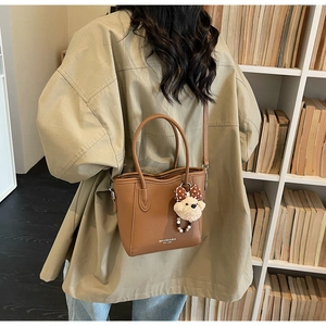 Brown lychee leather bucket bag with plush bear charm and brand logo against a bookshelf