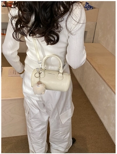 Chic cream cylinder handbag with a white plush charm shown in a lifestyle street setting
