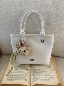 White faux leather tote bag with knot handle and teddy bear charm on textured background