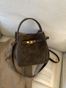 Brown suede bucket bag with gold lock on a beige sofa next to a book and trench coat