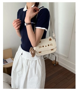 Cream cat-shaped crossbody bag with bear plush charm shown on model in sunny room