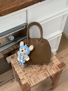 Brown lychee leather shell handbag with bear charm on a woven stool in a kitchen