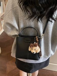 Stylish black leather bucket bag with plush dog charm shown in a crossbody wear style
