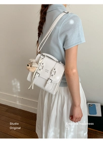 Detail of white square crossbody bag with buckle straps and a cute brown plaid teddy bear charm