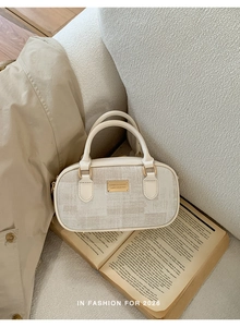 Détail d'un sac Boston crème avec logo doré sur canapé avec un livre décoratif