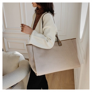 Model wearing a large beige canvas bucket tote bag with brown leather trim and chain strap