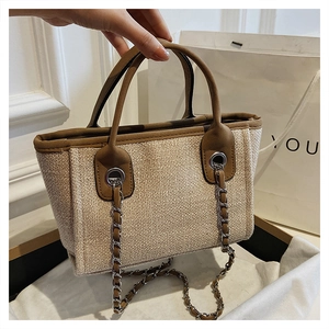 Detail of woven straw tote bag featuring brown leather trim and metal chain strap