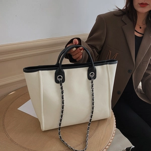 Minimalist beige canvas tote bag with black leather trim and chain strap on a wooden table