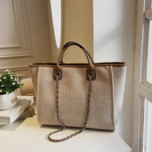 Large beige canvas bucket tote bag with brown leather handles and decorative metal chain