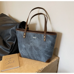 Detail of grey-blue tote bag with brown leather trim and golden metal bar on stone surface