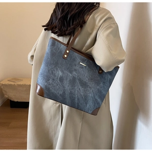 Detail of grey-blue tote bag with brown leather trim and metal accents on beige trench coat
