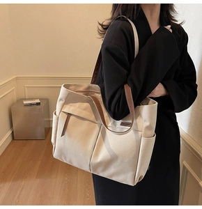 Model wearing a large beige canvas tote bag with brown leather handles in a minimal room