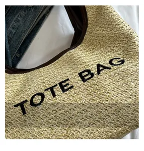 Detail of straw tote bag featuring black embroidered text and premium brown piping trim