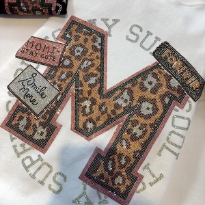Détail d'un t-shirt en coton blanc avec un M en strass léopard et des slogans brillants