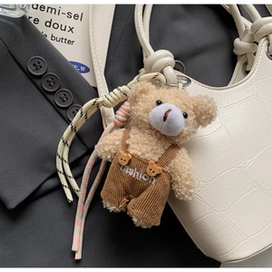 Detail of beige teddy bear charm with fashion overalls on a white textured shoulder bag