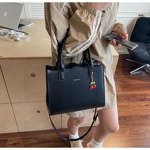 Detail of black leather tote bag with cherry charm and metal logo in indoor setting