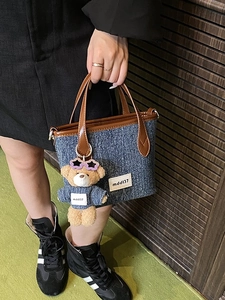 Denim bucket bag with brown leather trim and a cute teddy bear charm on a wood floor