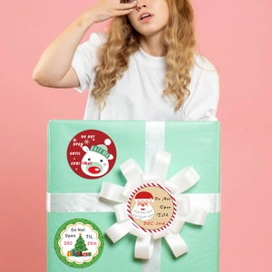 Three festive Christmas gift stickers on a large gift box held by a model with pink background