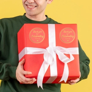 Pink and gold foil Thank You stickers on a red gift box held by a person in a main view