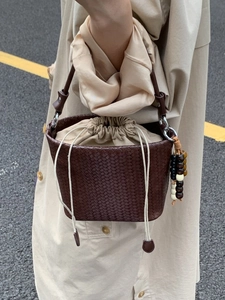 Brown woven bucket bag with drawstring liner and beaded charm held over a beige coat