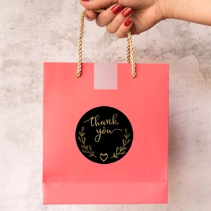 Detail of round black and gold thank you sticker on a pink paper shopping bag handle