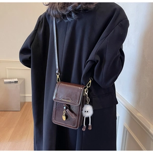 Detail of a shiny chocolate crossbody phone bag with a plush charm on a black coat