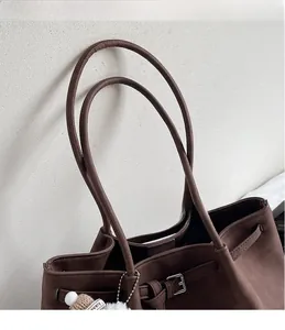 Close-up of a dark brown suede tote bag with long slim handles and a white fluffy charm
