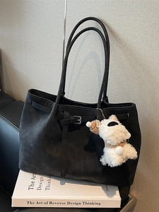 Minimalist black suede tote bag with silver buckle and plush puppy charm on a leather sofa