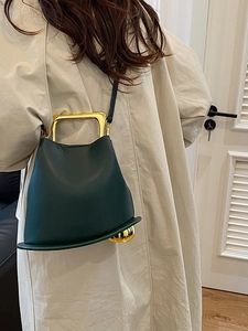 Dark green bucket bag with gold cat-ear handles shown as a crossbody on a beige coat