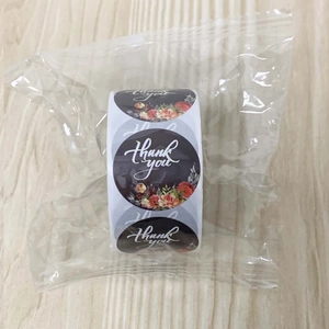 Close-up of floral Thank You stickers on roll with dark background and orange flowers