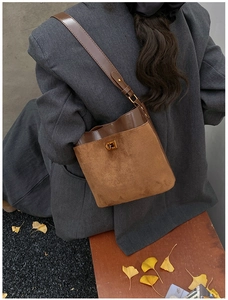 Brown suede bucket bag with gold buckle shown on shoulder against autumn leaves