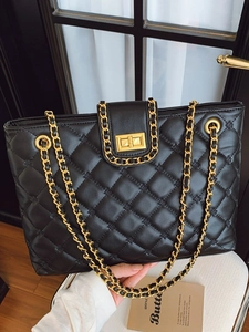 Chic black quilted tote bag with gold chain straps and turn-lock closure on white chair