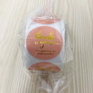 Close-up of a roll of gold foil Thank You stickers on a wood background with packaging