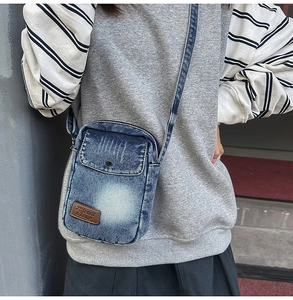 Detail of blue denim crossbody bag featuring metal snap button and logo patch on front