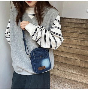 Detail view of a retro blue denim crossbody phone bag worn over grey casual outfit