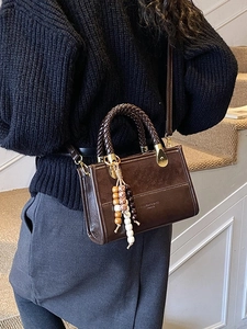 Brown box handbag with woven handle and beaded tassel shown on a stylish shoulder model