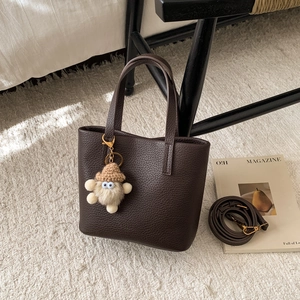 Detail of dark brown litchi grain leather tote bag with plush charm on a soft rug