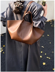 Brown leather wing-shaped tote bag shown in a street style scene with autumn leaves