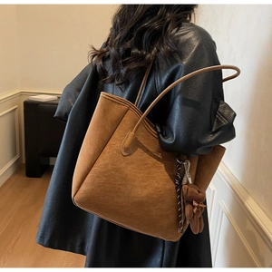 Brown suede tote bag with plush bear charm worn by a model in a cozy indoor setting